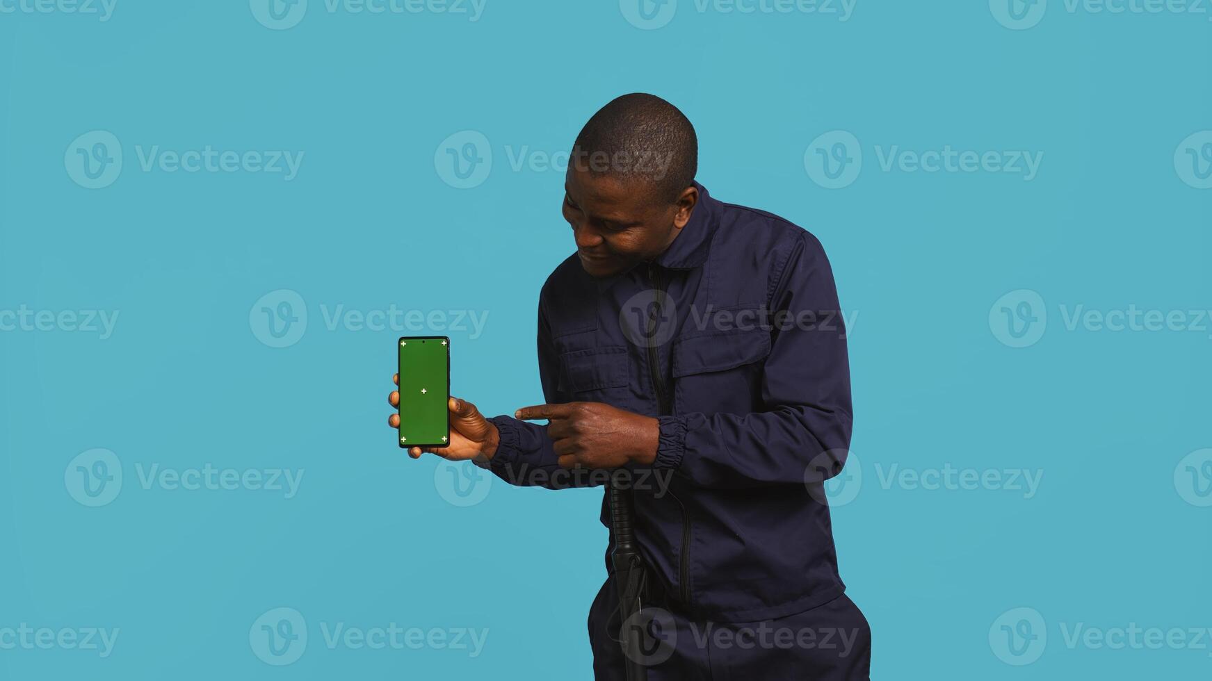 Portrait of joyous security guard at work showcasing green screen phone while overseeing premises. Jolly private law enforcement worker presenting mockup smartphone, studio backdrop, camera B photo