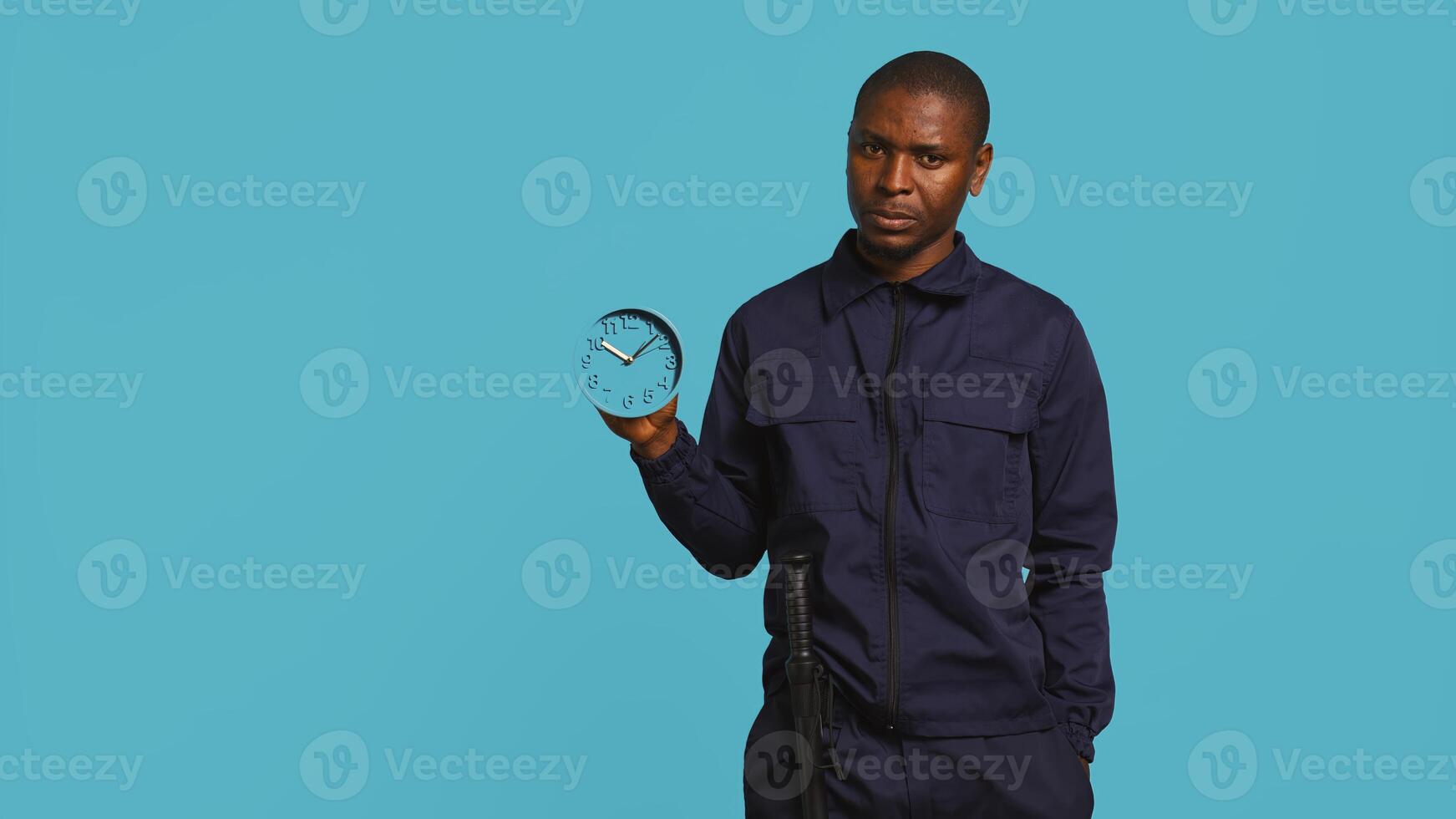 Security guard holding clock, rushing people out of property. Portrait of stern bodyguard showing visitors time is passing, preparing for closing down location, studio background, camera B photo