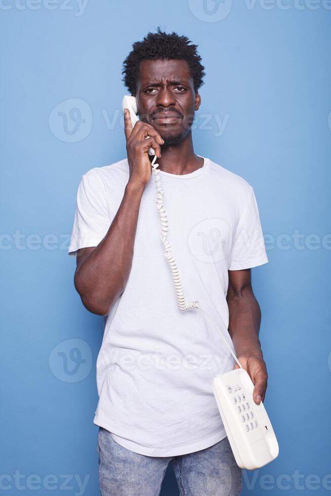 Black man in casual clothing speaking on his telephone against isolated blue backdrop. He looks surprised and confused, holding the landline close to his ear, with the phone cord visible. photo
