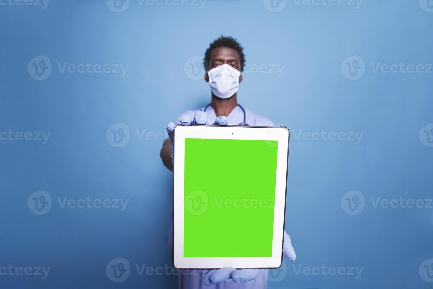 African American healthcare specialist in scrubs and face mask stands with tablet device showing green screen on blue background. Black man holding a tablet with copyspace mockup template. photo