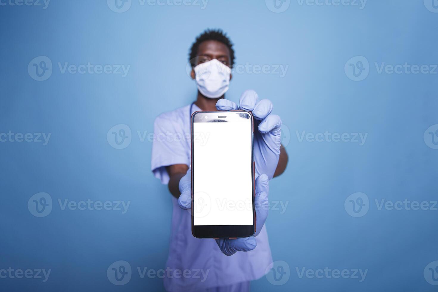 African American doctor stands confidently in scrubs, holding a mobile device with a blank white screen. Male medical assistant vertically holding smartphone with isolated copyspace mockup template. photo