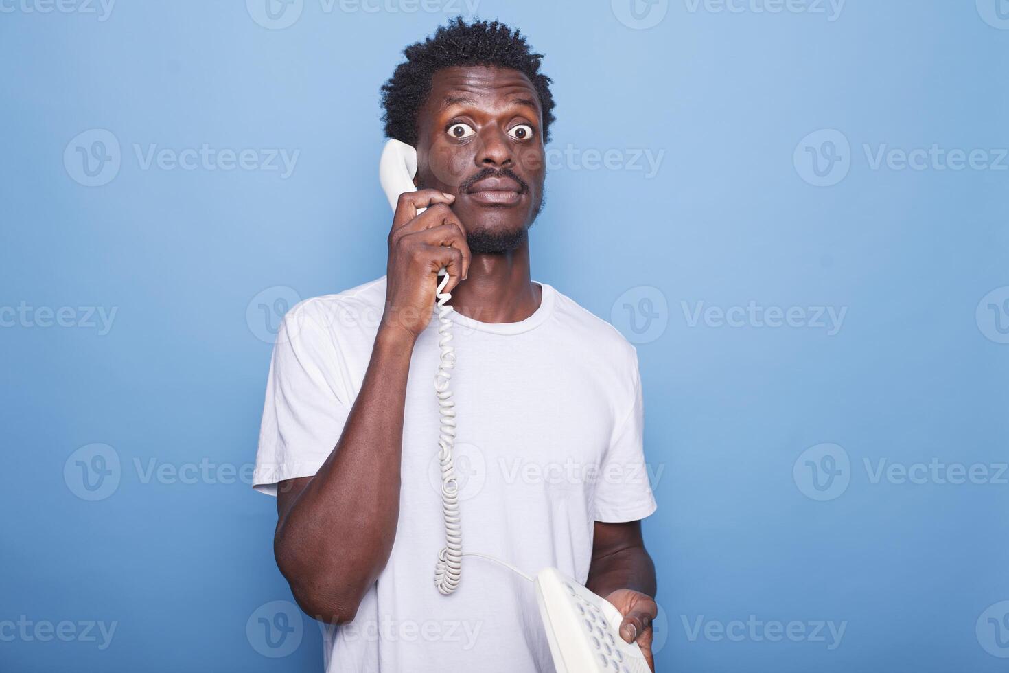 Surprised black man with afro hair on blue background talking on a landline phone. Portrait of shocked african american male individual communicating through the telephone. photo