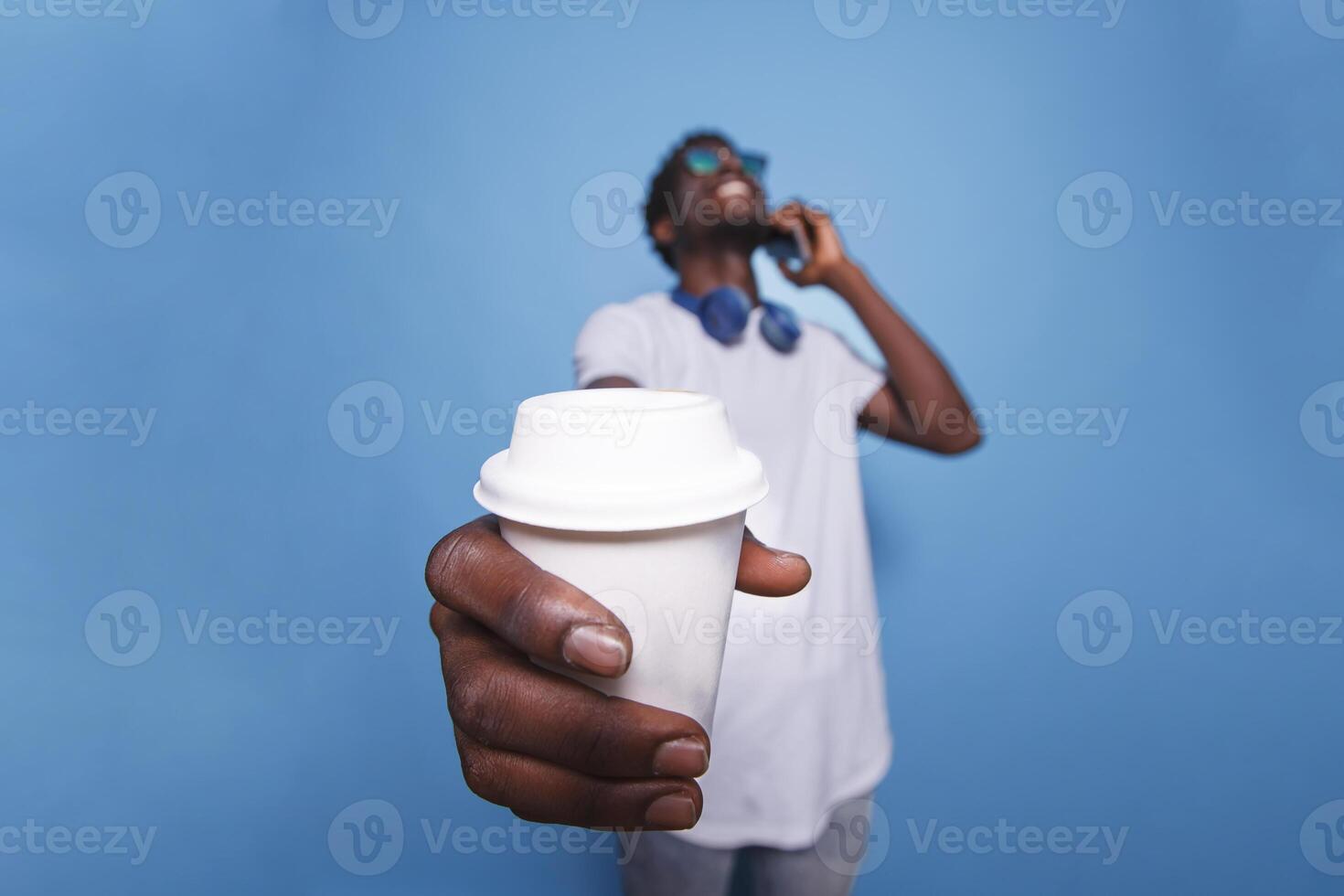 Black man laughing and grasping a coffee cup near the camera. Over an isolated background, a casual African American adult is on the phone, wearing sunglasses, and holding a beverage. photo