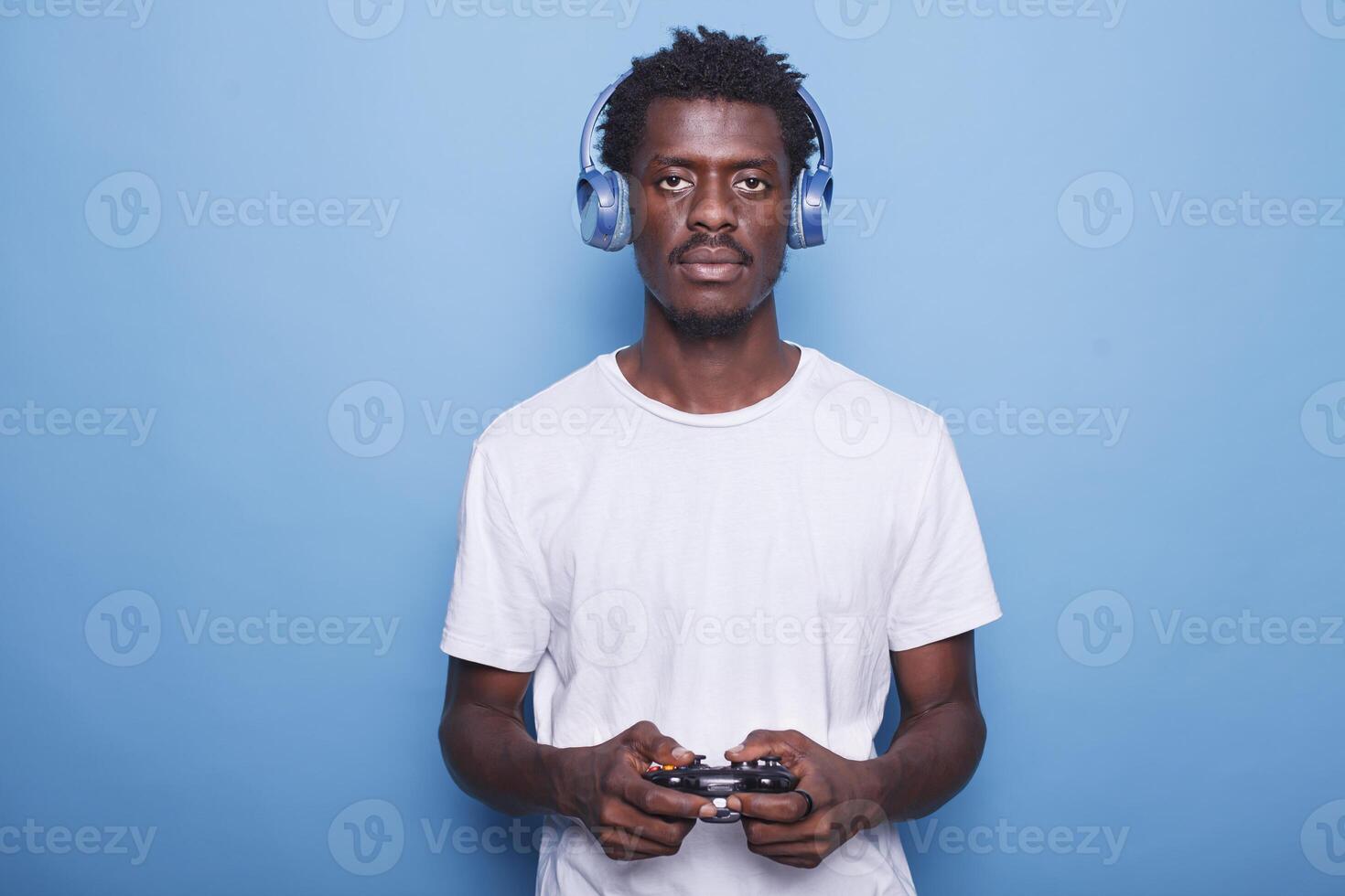 Black man uses headphones and wireless controller while staring at camera. African American guy using joystick to play games, enjoying virtual activities, and listening to music. photo