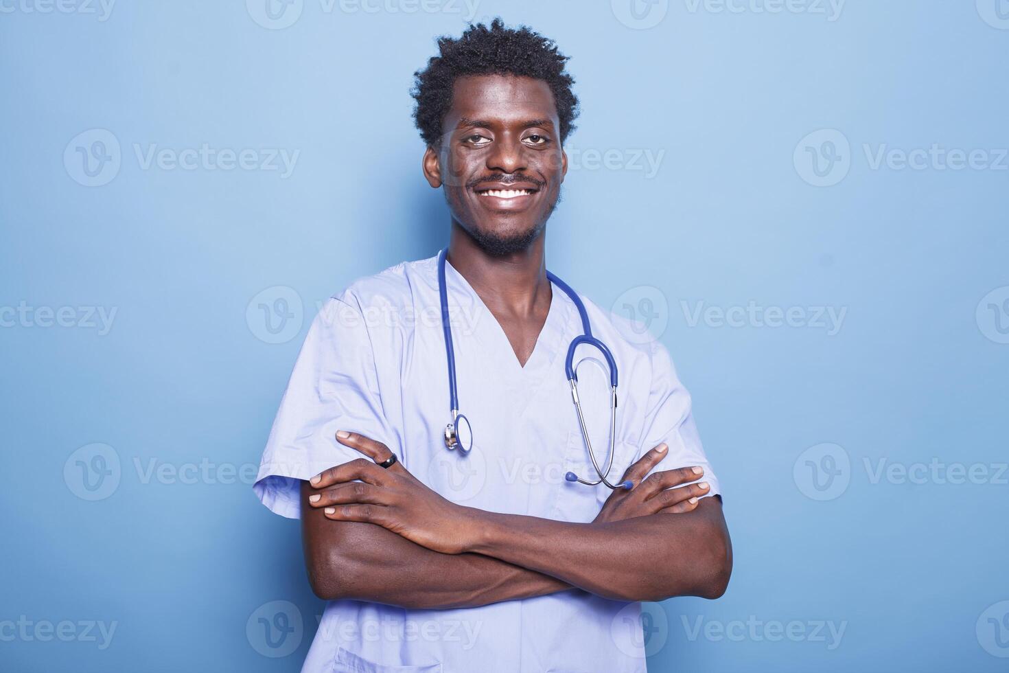 Smiling black man wearing blue scrubs and stethoscope posing in front of camera. Portrait of african american medical specialist standing against isolated background with arms crossed. photo