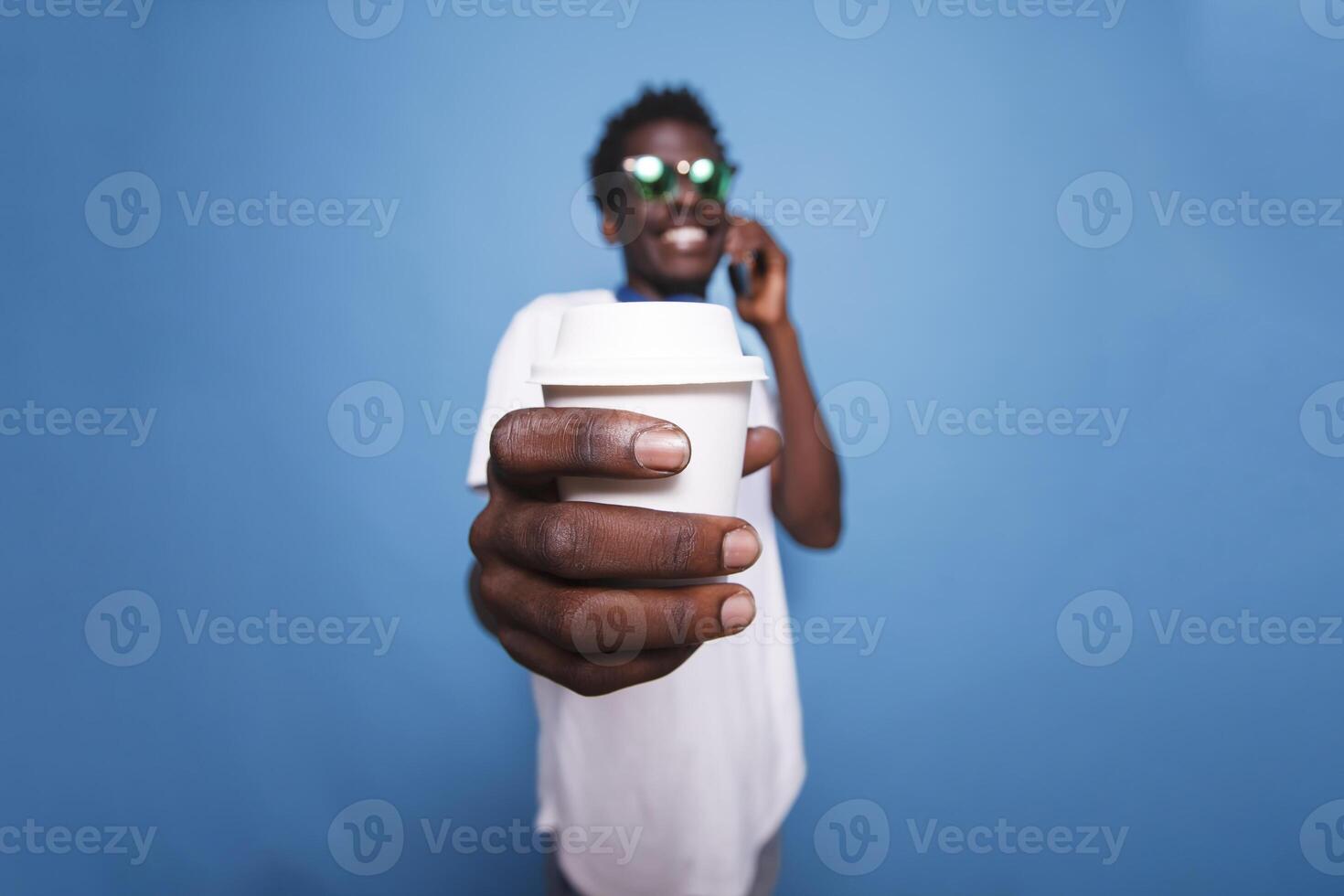 Near the camera, black man is laughing and gripping a coffee cup. Casual African American adult is on the phone over an isolated background, wearing sunglasses and holding a beverage. photo