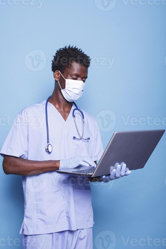 African american man with face mask standing and holding modern laptop. Medical assistant with blue uniform and stethoscope using personal computer in front of isolated background. photo