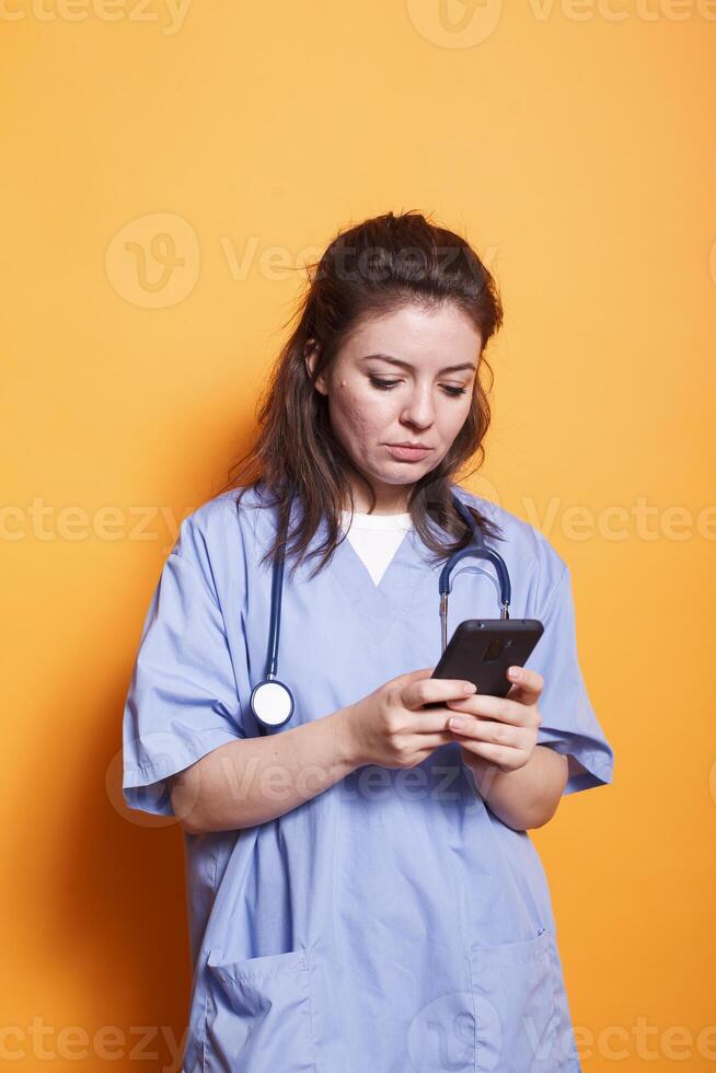 Portrait of nurse practitioner with stethoscope surfing the net on her mobile device. Female doctor wearing blue scrubs is using cell phone for communication with hospital coworkers. photo