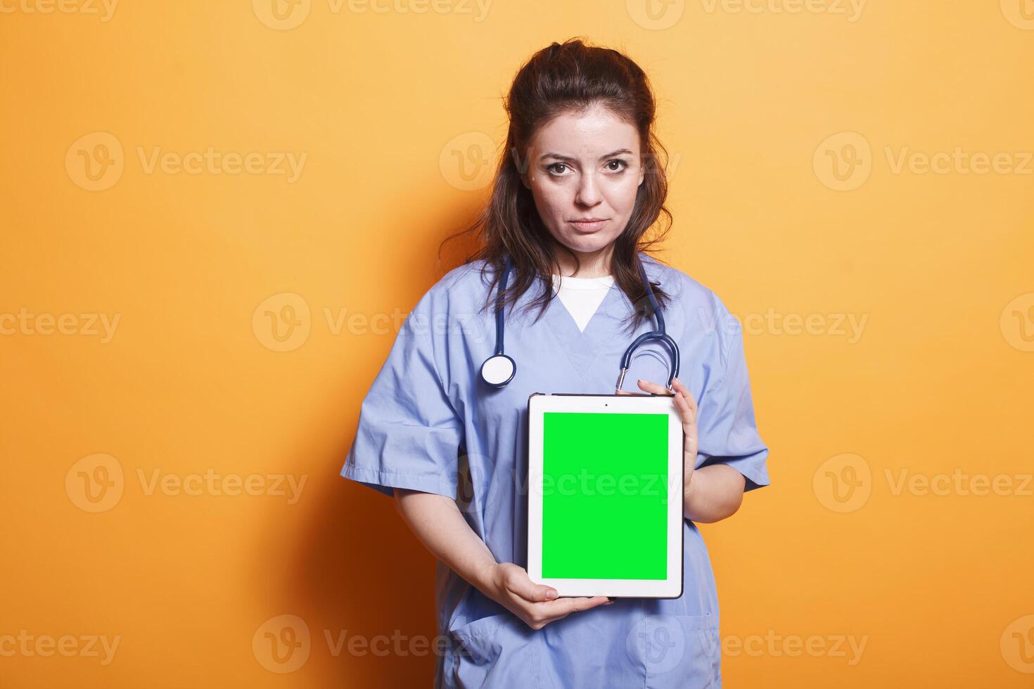 Healthcare professional vertically holding tablet with green screen. Woman wearing nurse uniform and stethoscope is grasping digital device displaying blank chromakey mockup template. photo
