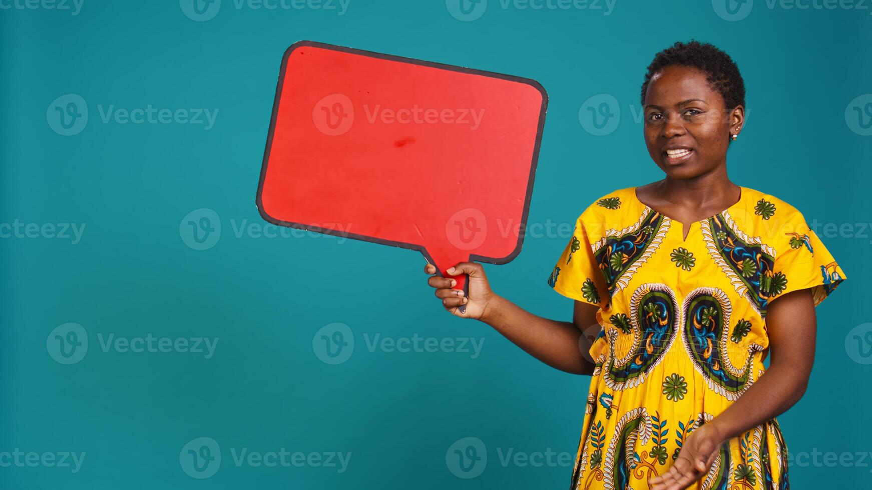 Smiling person showing a red isolated speech bubble in studio, holding a cardboard icon for a commercial advertisement. Young woman in tribal attire presenting an empty carton sign. Camera B. photo
