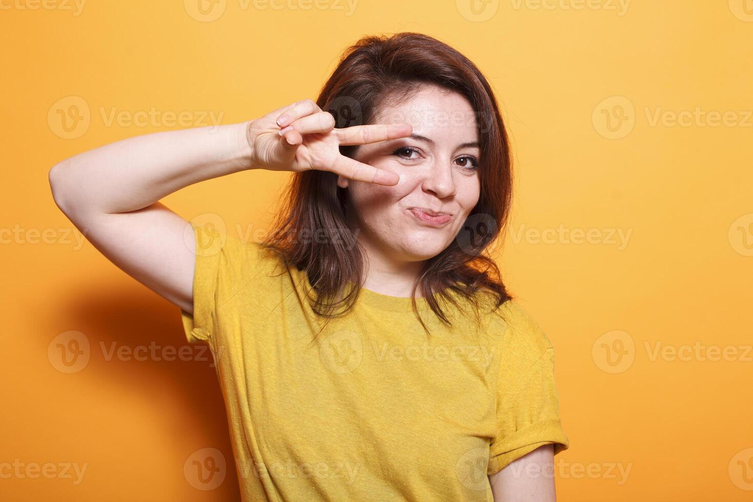 Cheerful brunette with bright smile and casual outfit stands out against isolated background in this waist-up portrait. Carefree lady holding up two fingers to symbolize peace, feeling upbeat. photo