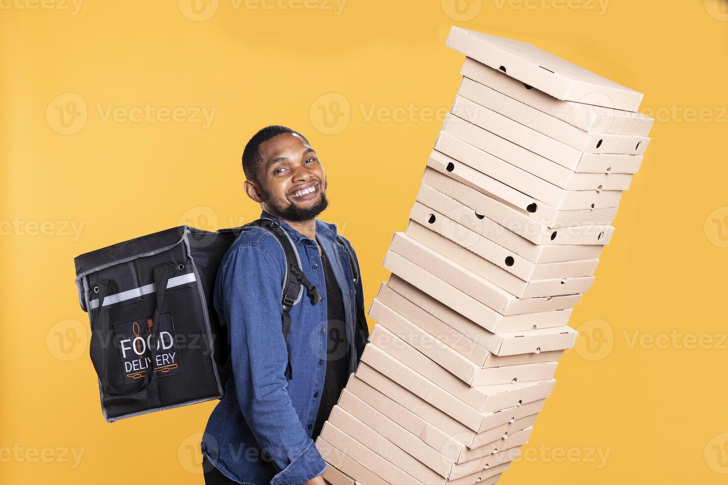 Smiling deliveryman struggles to hold a huge stack of pizza boxes in studio, working on food delivery service to bring orders to clients. Express courier with a backpack carries stack of takeout. photo