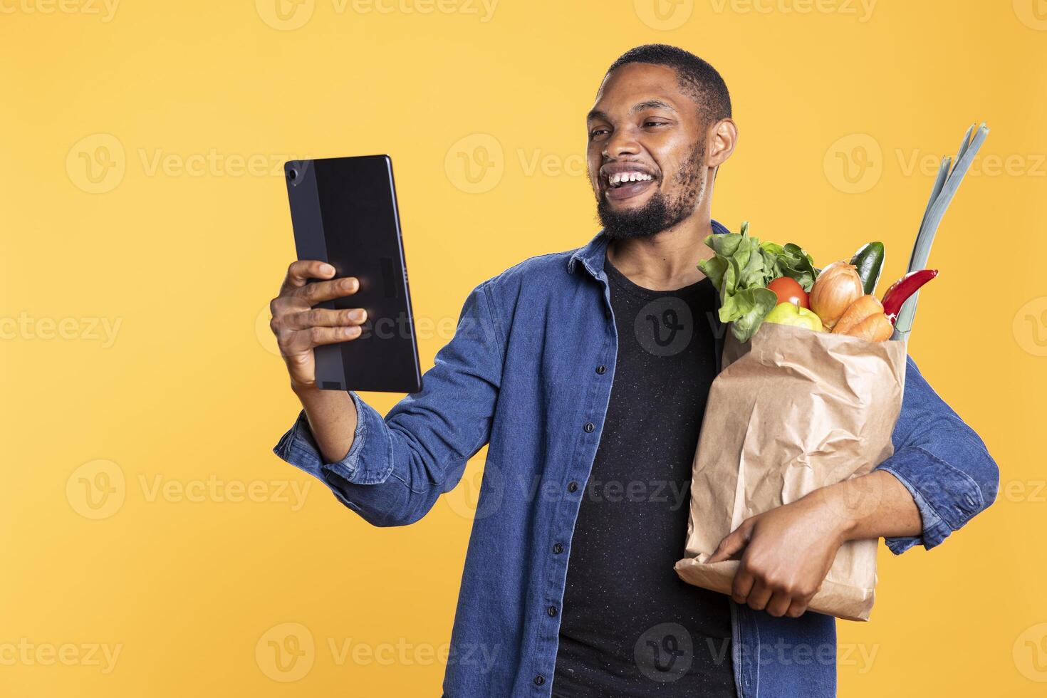 Smiling young man checks something on his tablet while he is holding a bag full of fresh vegetables and groceries. Confident guy uses his mobile device for social media against yellow backdrop. photo