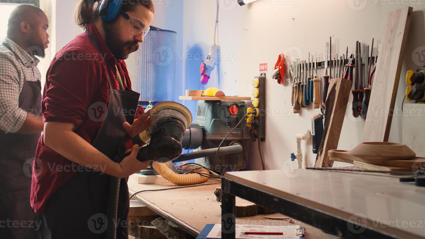 Carpenter at work consulting blueprint, using orbital sander on lumber, wearing safety glasses. Artisan with protecting equipment uses angle grinder on wood after checking schematics, camera B photo