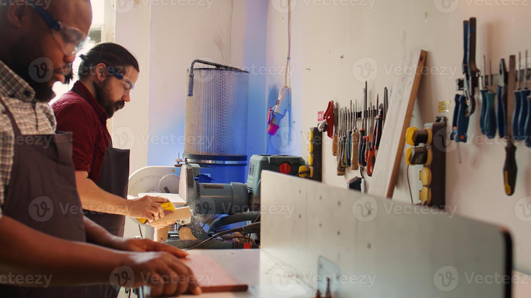 Team of woodworkers in assembly shop using sandpaper and spindle moulder for sanding wooden surface before painting it. Carpenters wearing safety glasses using abrasive sponges on wood, camera B photo