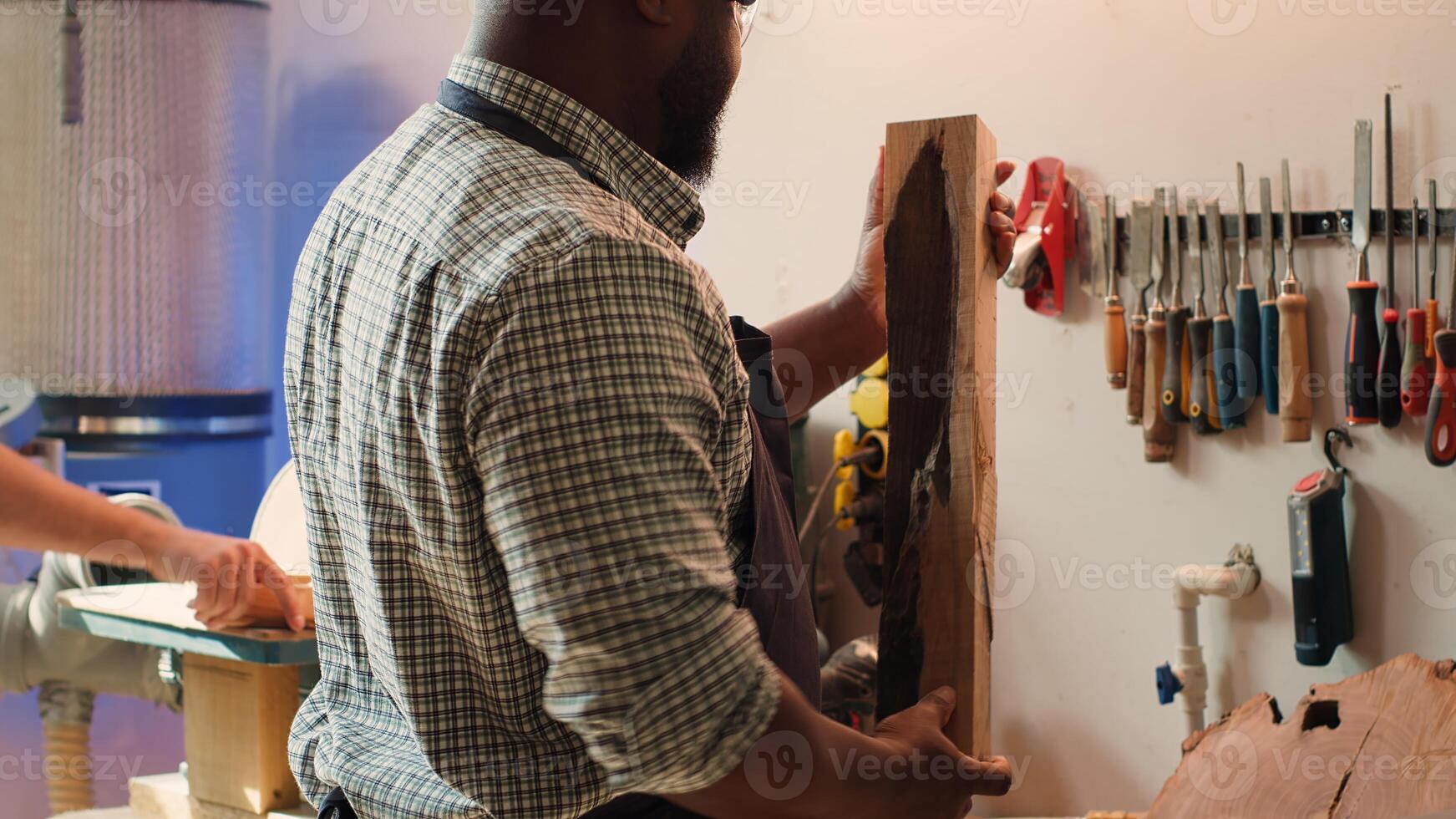 BIPOC carpenter in woodworking shop inspecting lumber piece before assembling furniture, checking for scratches. Cabinetmaker evaluates timber block, picking right tool from rack for it, camera A photo