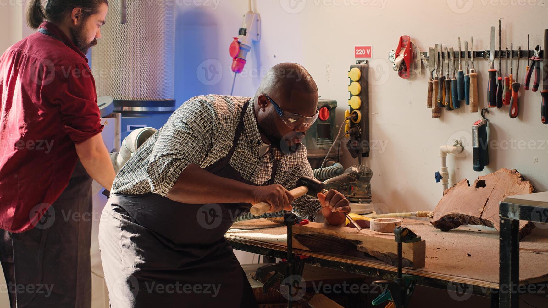 Sculptor shaping raw timber using chisel and hammer in carpentry shop with coworker cleaning workbench in background. BIPOC man making wood sculptures, engraving plank with tools, camera A photo