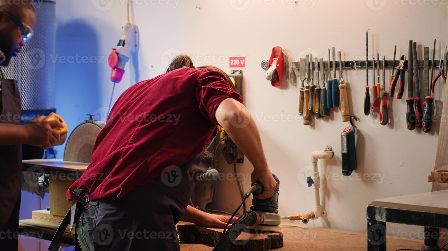 Carpenter collaborating with apprentice, using orbital sander with fine sandpaper to achieve refined finish. Manufacturer and BIPOC colleague using angle grinder on wood block, camera A photo
