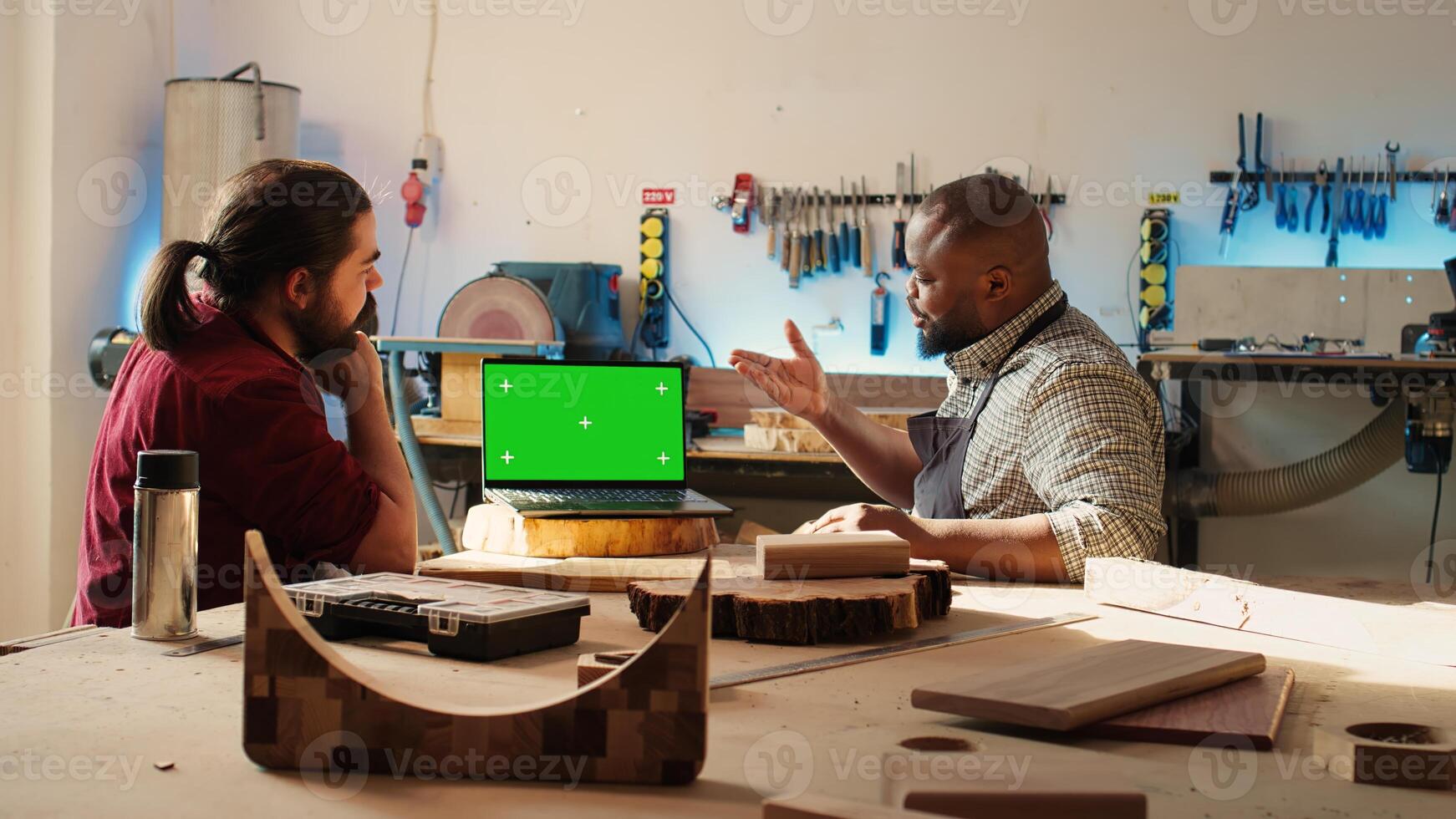 Joyous cabinetmaker looks over blueprints schematics on isolated screen laptop in CAD software with apprentice. Smiling artisan and colleague designing custom 3D furniture on mockup notebook, camera B photo