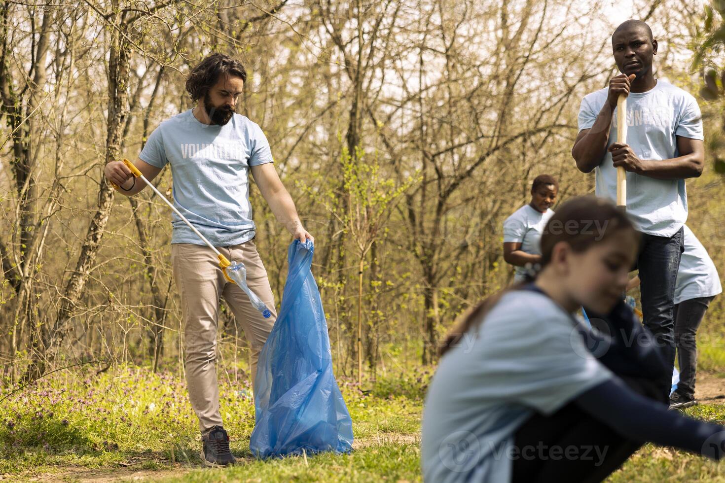 Diverse activists picking up garbage and plastic trash with tongs, working to fix illegal dumping and protect natural forest ecosystem. People volunteering for litter cleanup mission. photo