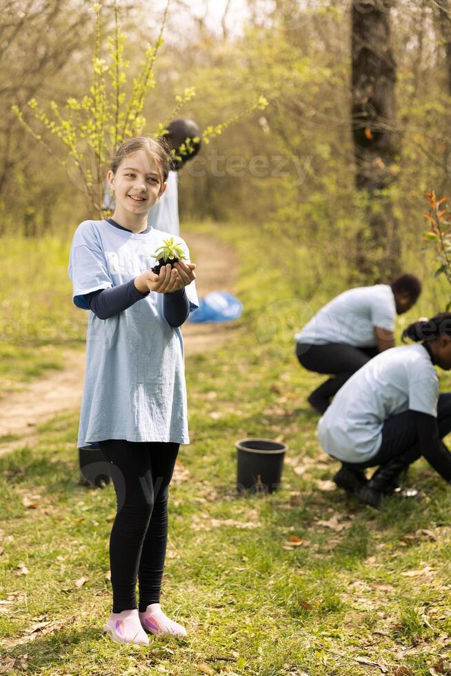 Little girl activist showing small seedling with natural soil, presenting a green plant for new habitat in the forest. Child fighting nature preservation, cultivating consciousness and awareness. photo