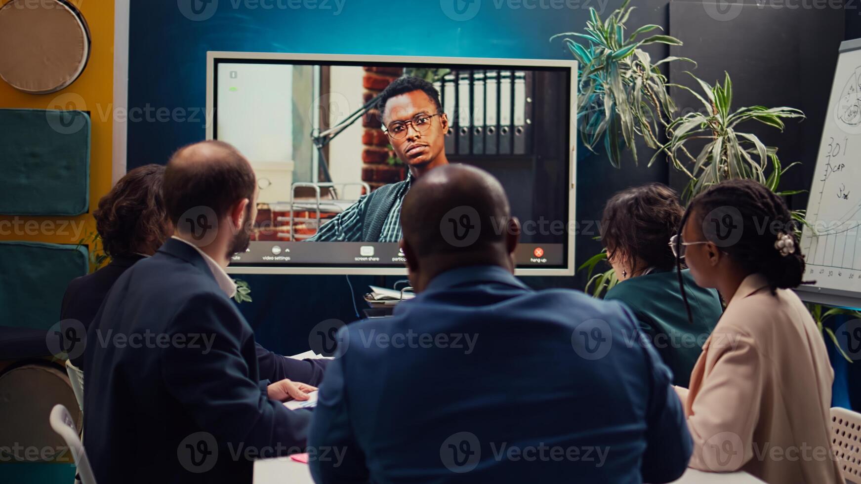 Management team meeting with CEO on videocall connection in a boardroom, briefing him about market trends and project resource allocation. Employees discuss with the director. Camera B. photo