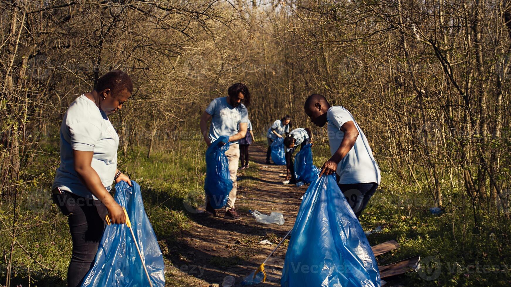 Diverse activists cleaning up rubbish in a garbage disposal bag, environmental conservation concept. Volunteers protecting the forest ecosystem, collecting rubbish with claw tool. Camera B. photo