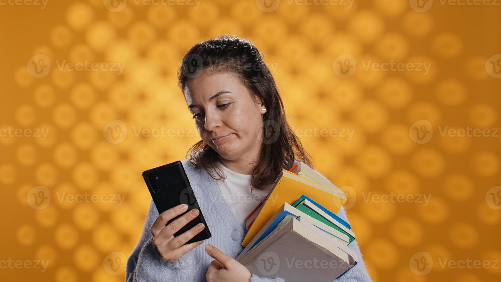 Upset woman holding stack of book, preparing for school exam using phone, studio background. Student with pile of textbooks pouting while researching for university homework using smartphone, camera B photo