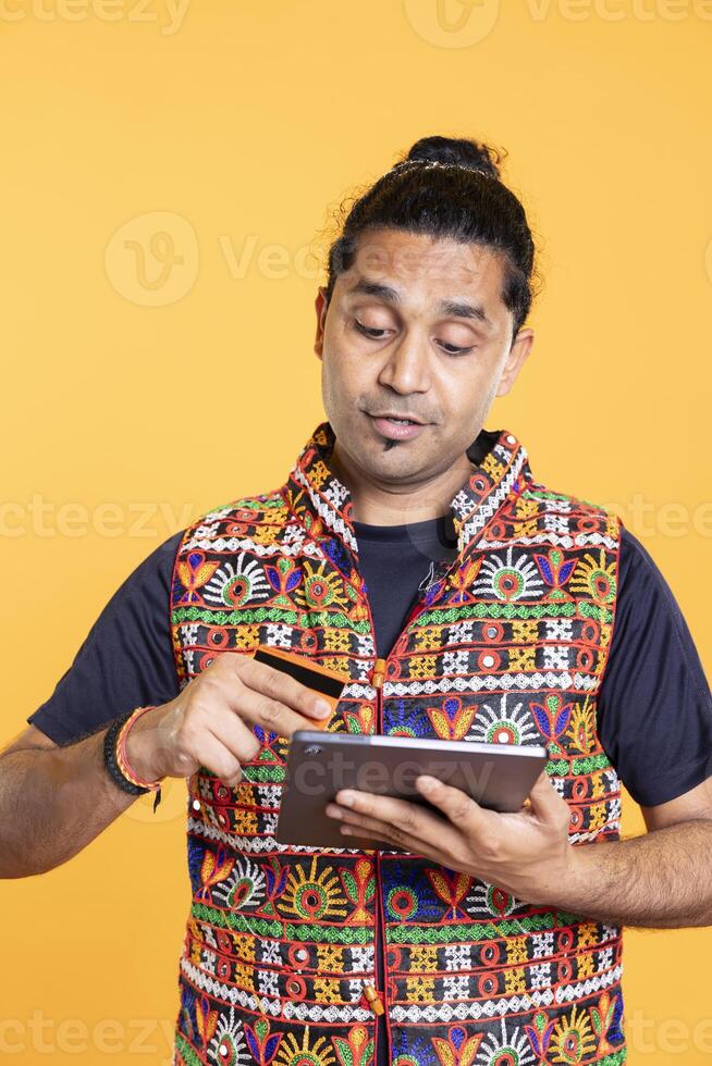 Man adding payment method on shopping website using tablet, taking advantage of promotions, studio background. Indian person buying stuff online using credit card, typing data on device photo