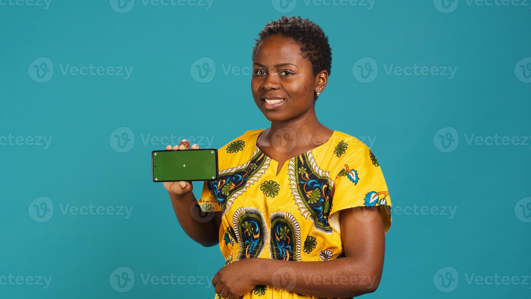 Woman presenting her smartphone with isolated copy space screen, creating an advertisement while she wears african traditional attire with tribal elements. Gorgeous young adult in studio. Camera A. photo