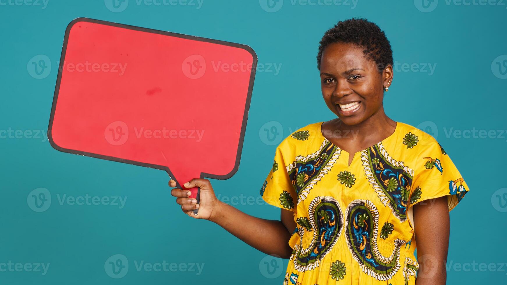 Smiling person showing a red isolated speech bubble in studio, holding a cardboard icon for a commercial advertisement. Young woman in tribal attire presenting an empty carton sign. Camera A. photo