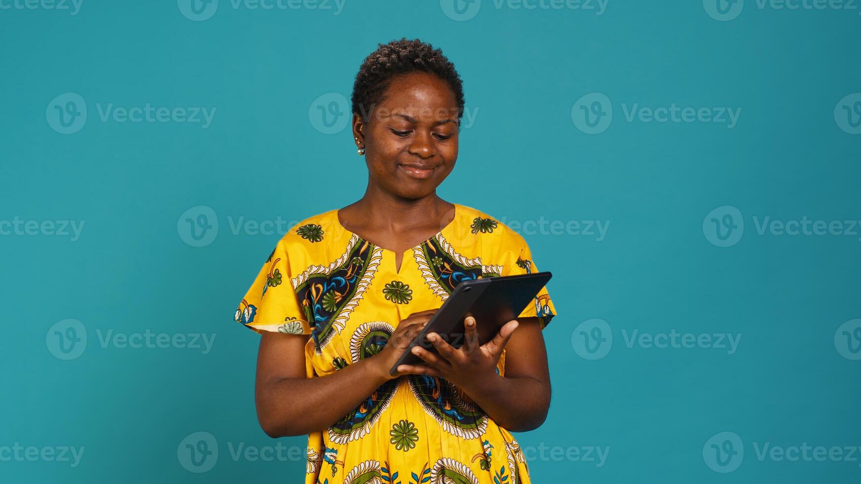 Smiling happy person checking internet websites on her tablet in studio, wearing african ethnic clothes with tribal elements. Confident pleased girl browses online social media sites. Camera A. photo