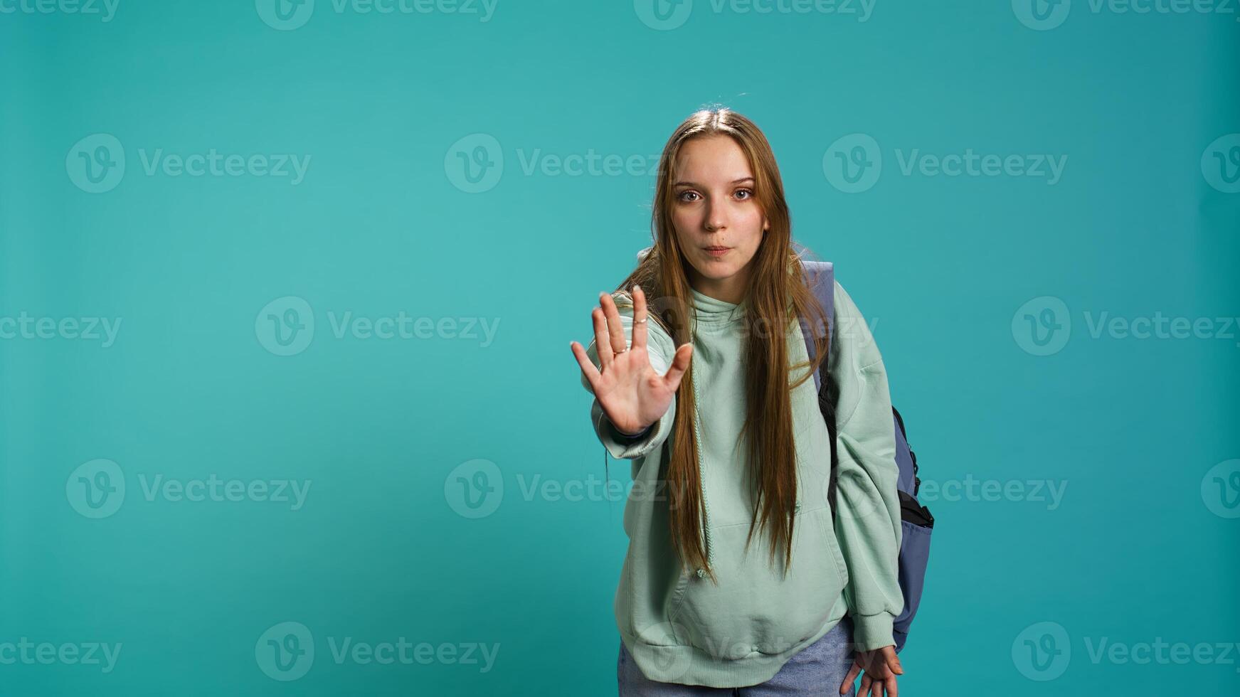 Assertive young woman doing stop hand gesture and talking. Annoyed girl doing firm halt sign gesturing, wishing to end concept, isolated over blue studio background, camera B photo