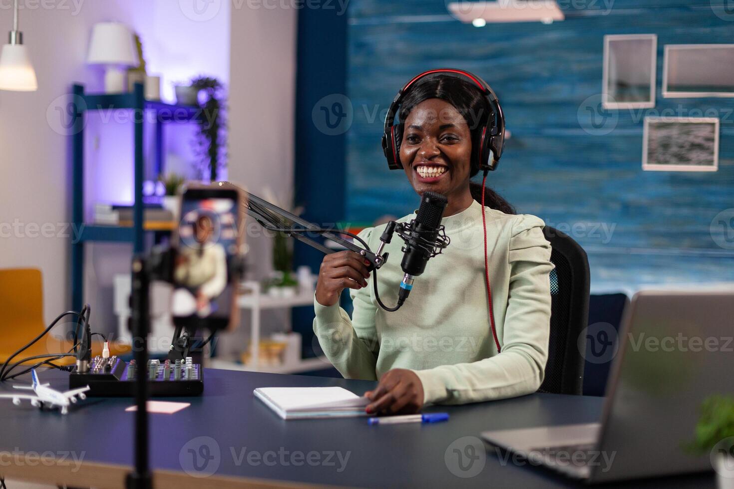 Podcast creator woman with headphones talking into production microphone answering subscribers questions filming content using smartphone in vlogging studio at home. Live stream photo