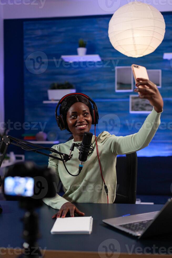 Woman influencer with headphones holding smartphone taking photo while recording vlog content using vlogging production equipment. African american social media vlogger hosting on air talk show