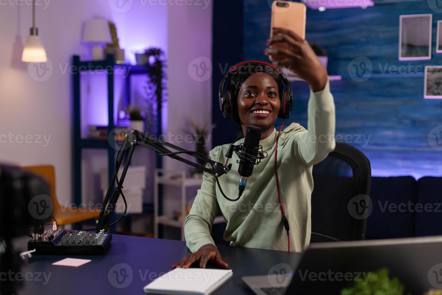 Social media influencer holding smartphone taking selfie while recording vlog content using vlogging production equipment. African american vlogger woman hosting on air talk show photo