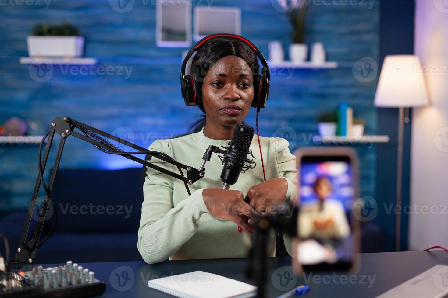 Social media influencer with headphones talking into production microphone answering subscribers questions filming content using smartphone in vlogging studio at home. Live stream photo