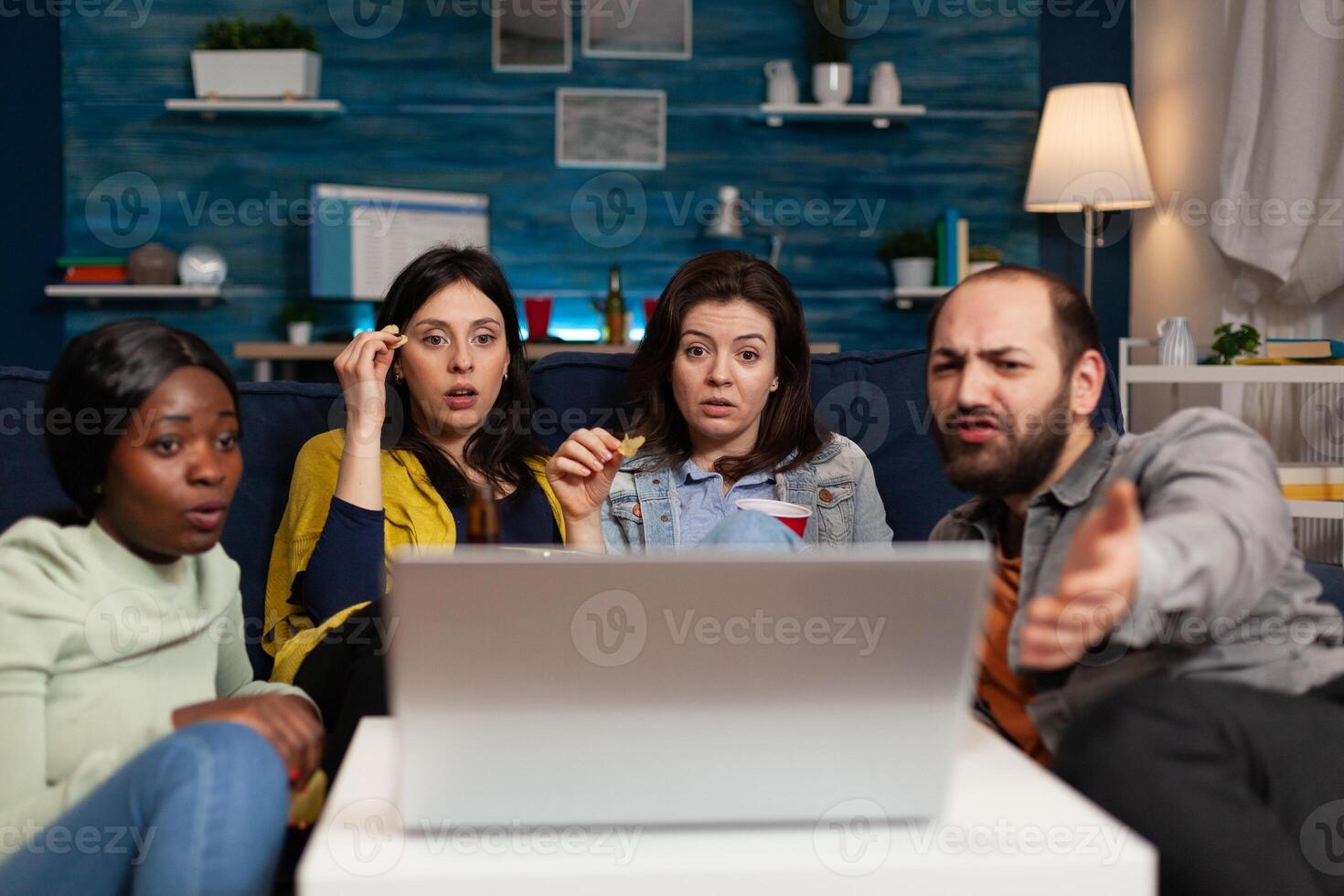 Portrait of multi-ethnic friends sitting on couch watching online movie on laptop computer enjoying hanging out together. Group of people enjoying relaxing time in living room. Friendship concept photo