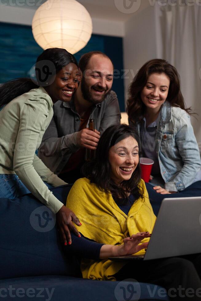 Multi-ethnic friends relaxing on couch laughting while watching fuuny comedy film during movie night at home enjoying spending time together. Group of people hanging out in evening. Friendship concept photo