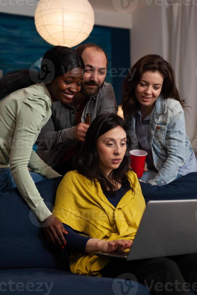 Multi-ethnic friends smiling while watching online comedy series on laptop computer during entertainment movie home night. Group of people enjoying hanging out together. Friendship concept photo