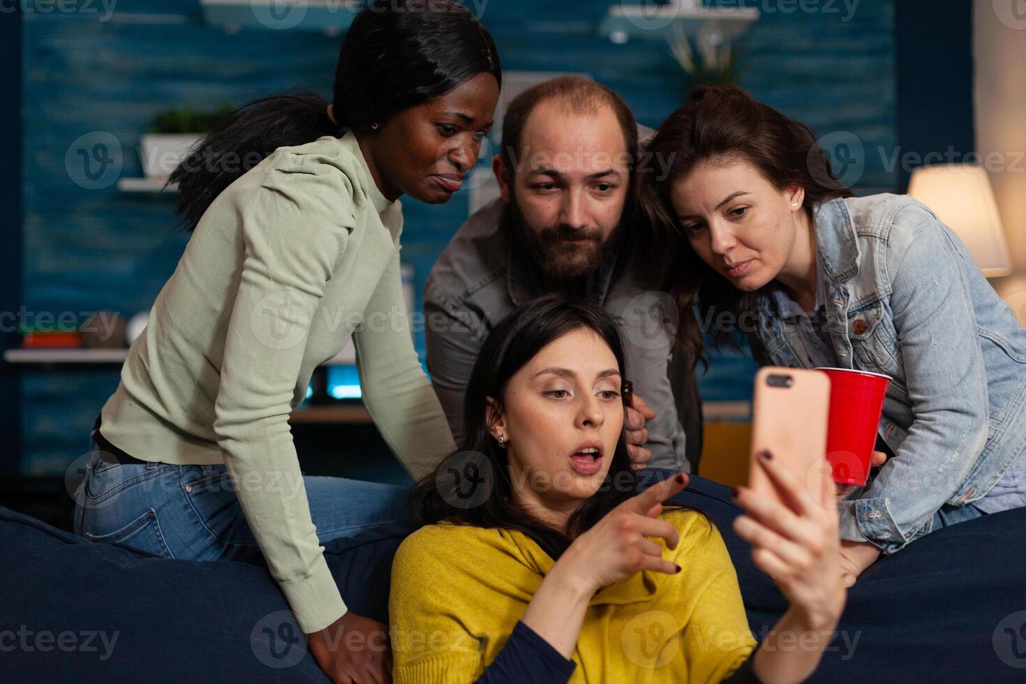 Multi-ethnic friends looking at fuuny videos on internet using smartphone during relaxing night at home enjoying hanging out together. Group of people having fun celebrating weekend in living room photo