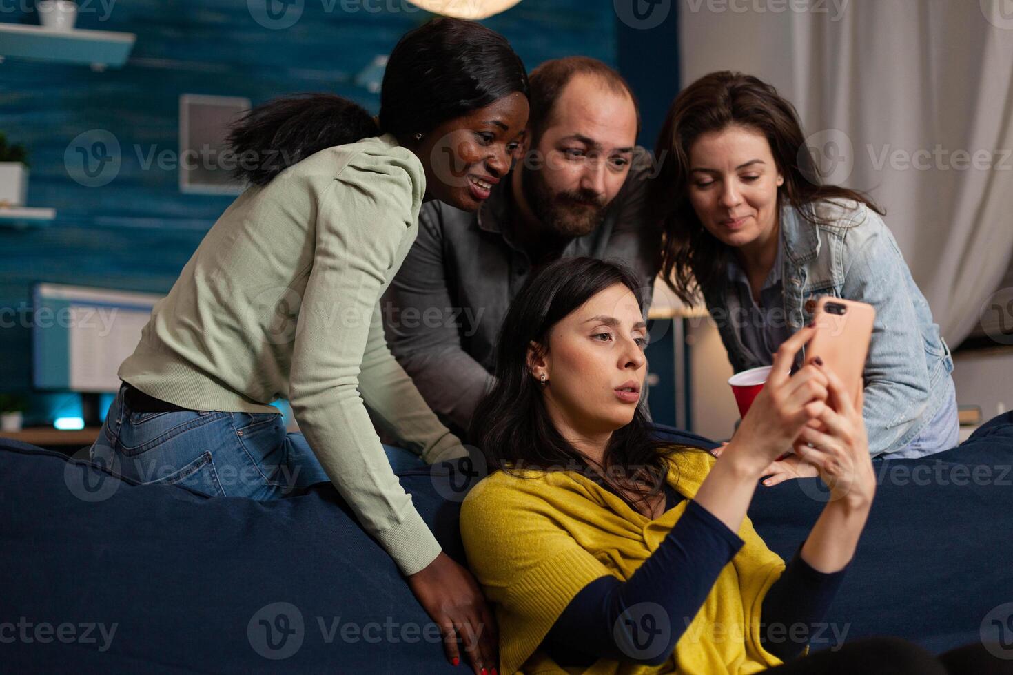 Multi-ethnic friends looking at fuuny videos on smartphone during movie night at home enjoying hanging out together. Group of people chilling in living room having fun. Friendship concept photo