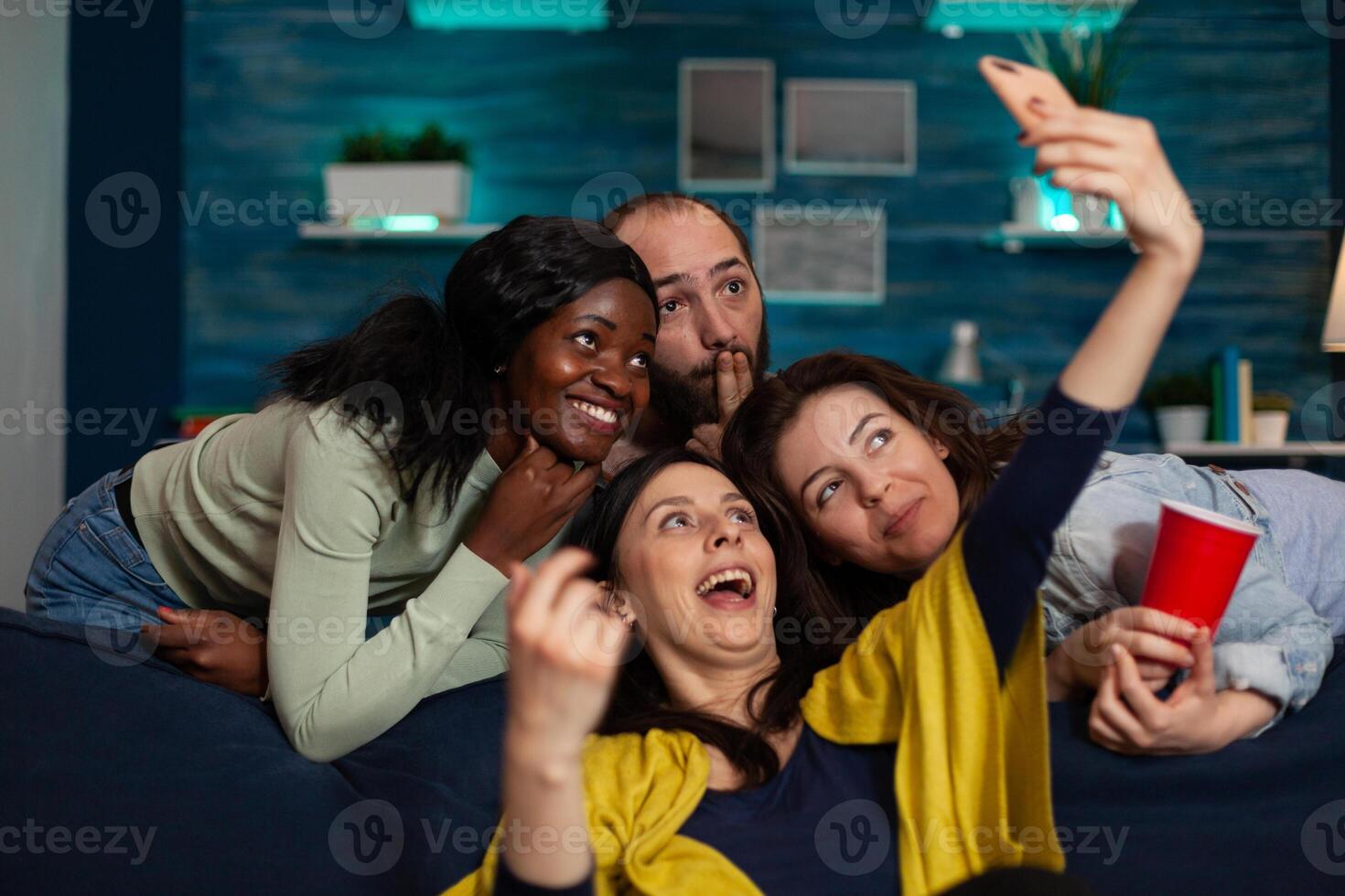 Multi-ethnic friends smiling at camera while smiling woman taking selfie sharing photos on social media celebrating wekeend at home. Group of people making funny expressio. Hanging out concept