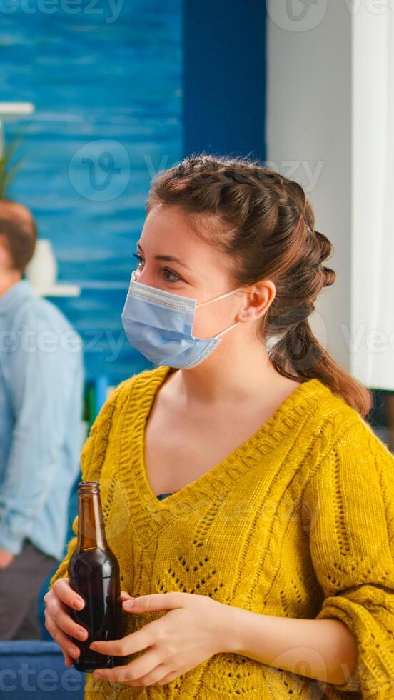 Diverse ethnic friends spending time together in apartment living room talking keeping social distancing wearing face mask, as prevention for coronavirus spreading in the course of global pandemic. photo