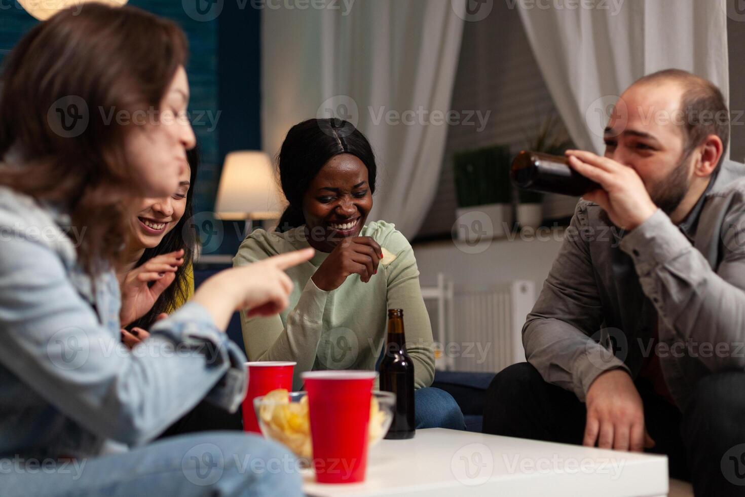 Multi-ethnic group of people enjoying spending time together laughing during wekeend party sitting on sofa in living room. Friends drinking beer, eating snack. Hanging out concept photo