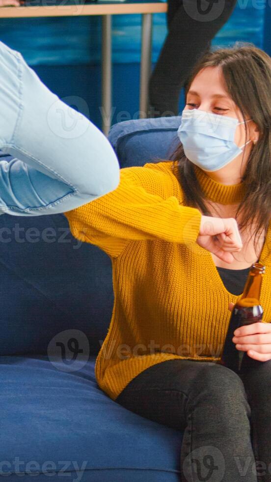 Happy cheerful friends touching elbow keeping social distancing greeting each other wearing face mask preventing spread of coronavirus, holding beer bottles socializing in apartment living room photo