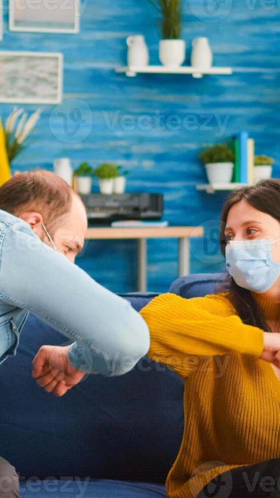 Friends with protection mask socializing during party sitting on sofa touching elbow keeping social distancing greeting each other preventing spread of coronavirus, holding beer bottles in living room photo