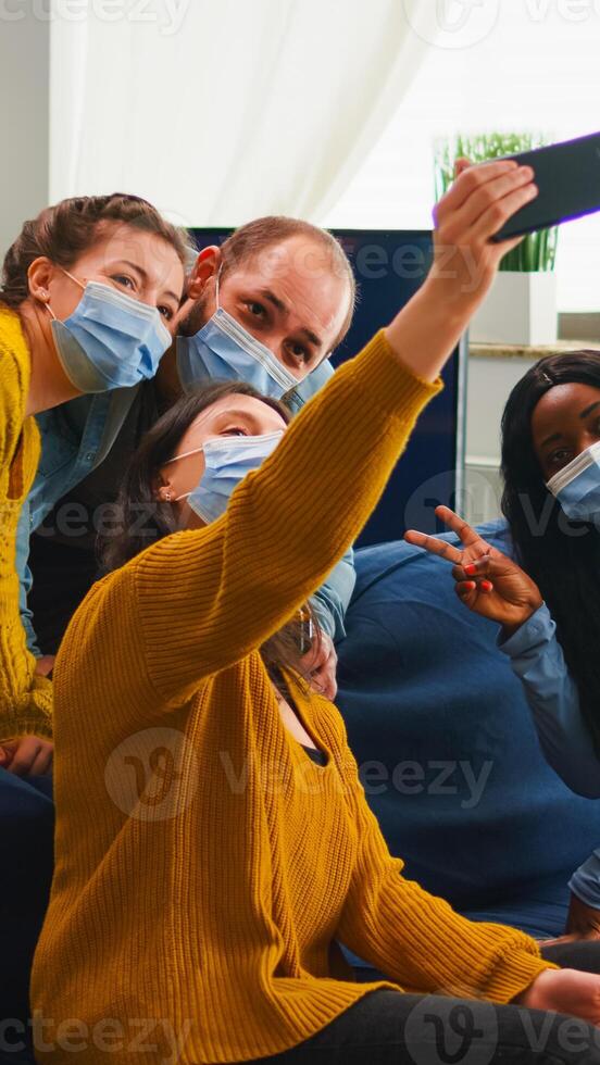 Multiracial friends taking selfie with face masks during covid 19 outbreak, new normal lifestyle concept with people having fun in living room respecting social distance to prevent virus spread photo