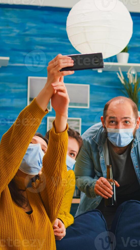 Cheerful happy mixed race friends taking selfie wearing face mask keeping social distancing against spread of virus sitting on couch in living room. Diverse people socializing keeping social distance photo
