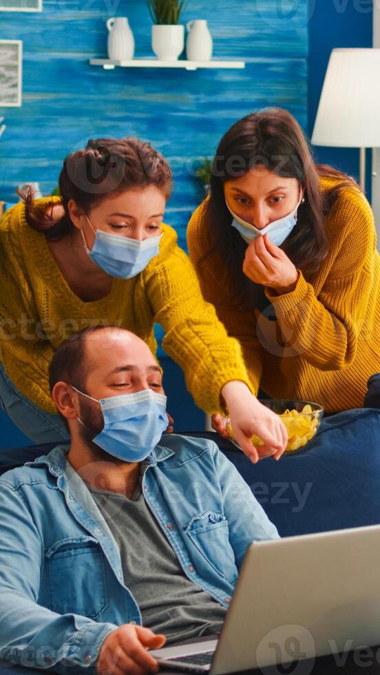 Cheerful happy multi ethnic friends looking at photo on laptop having fun together keeping social distance wearing face mask preventing coronavirus spreading. Diverse people enjoying new normal party